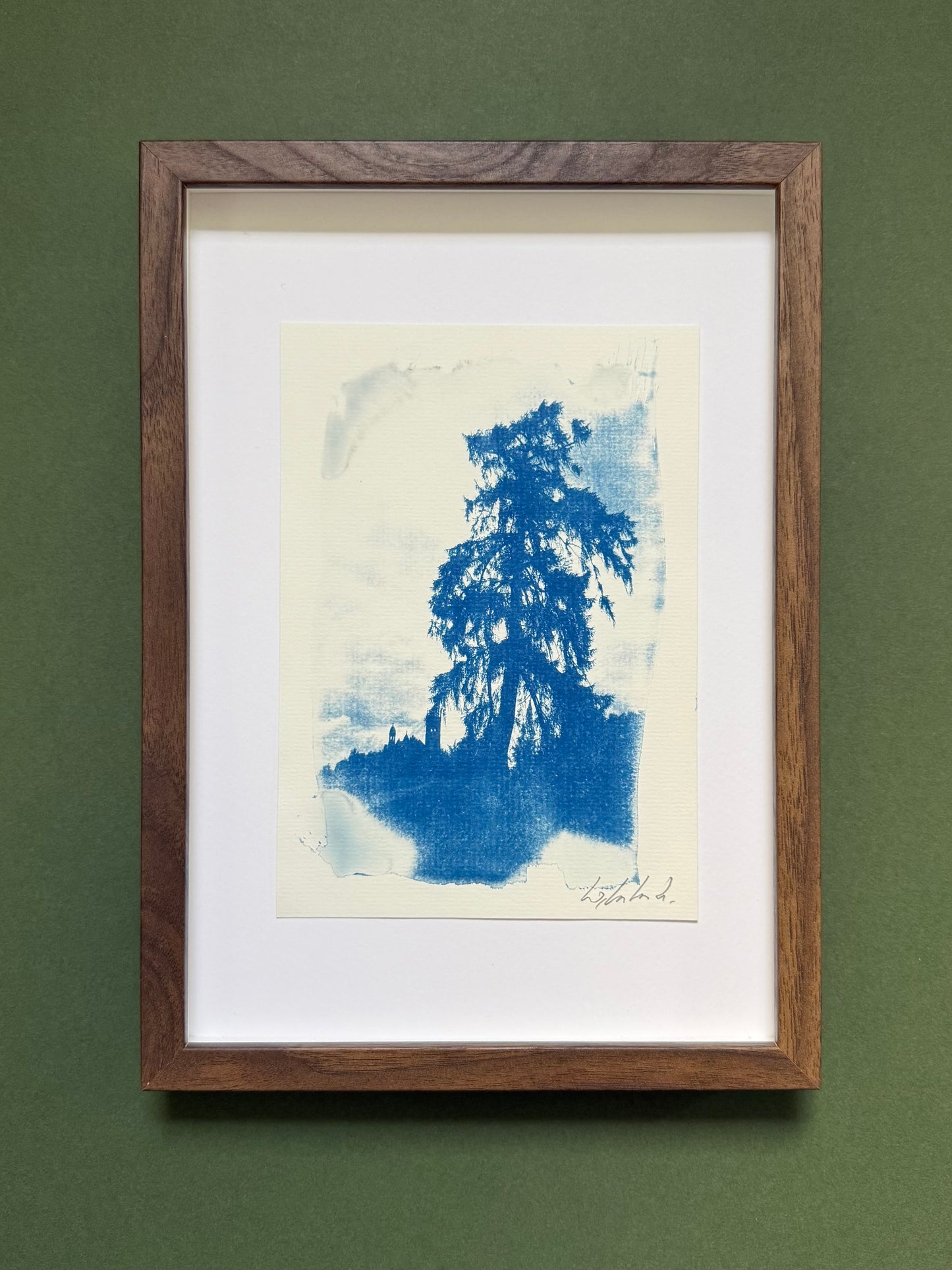 A framed blue ink tree artwork with a white mat and a medium-brown wooden frame, displayed against a solid dark green wall.