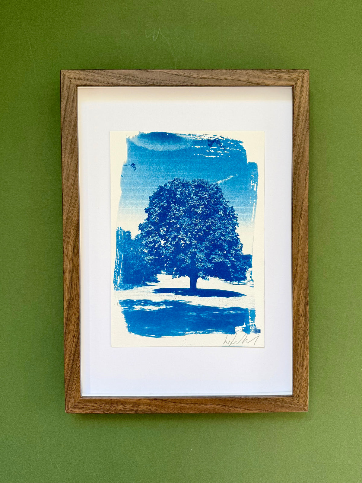 A framed art print featuring a blue ink linocut Cyanotype mounted on a white mat board and enclosed in a natural oak wood frame, displayed against a textured olive green wall.