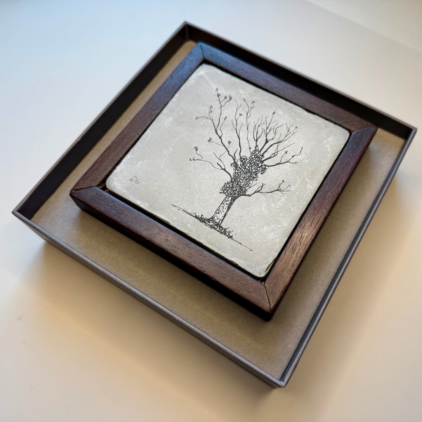 Framed cement tile artwork displayed at an angle inside a presentation box, showing the depth of the wooden frame and hand-drawn winter tree in ink.