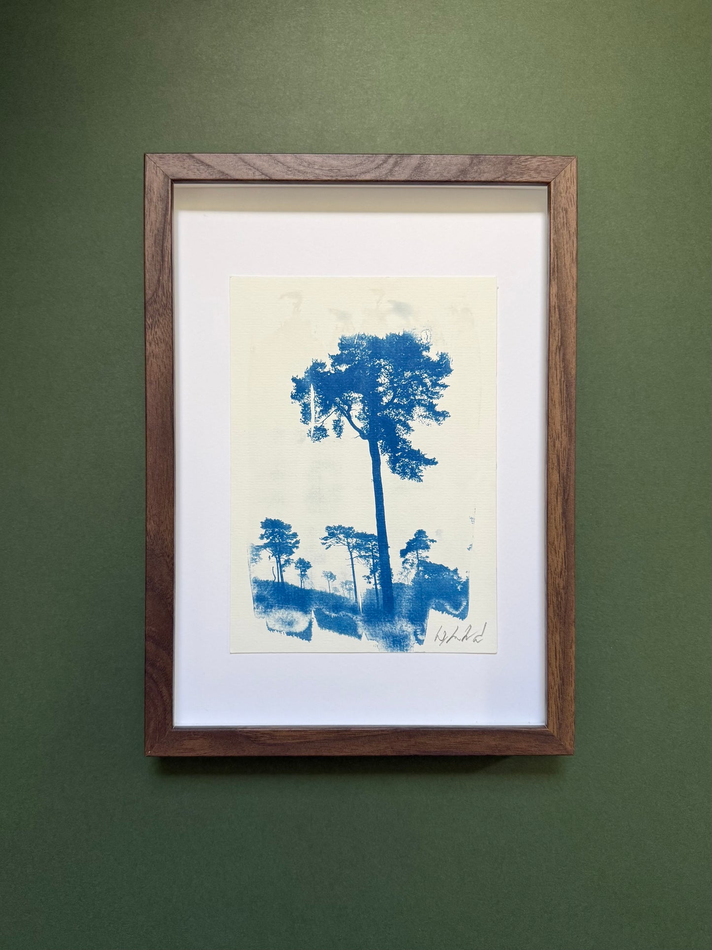 A framed blue ink tree landscape print with a white mat and a dark brown wood frame, mounted on a textured dark green wall.