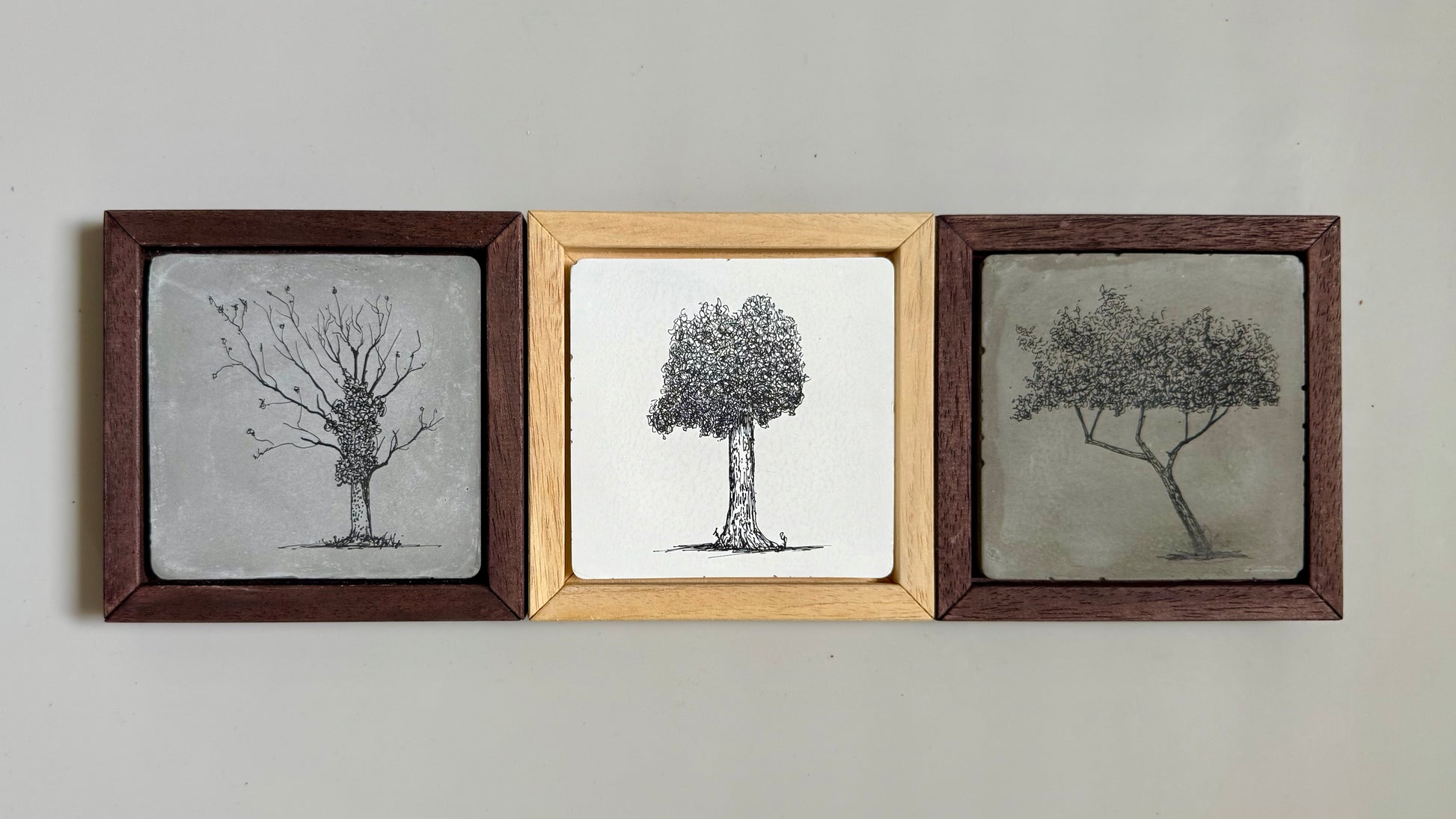 “Set of three framed ink tree drawings by Warren Garland. Each drawn on hand-cast cement tile with varied tree forms and wood frames in warm natural tones.”