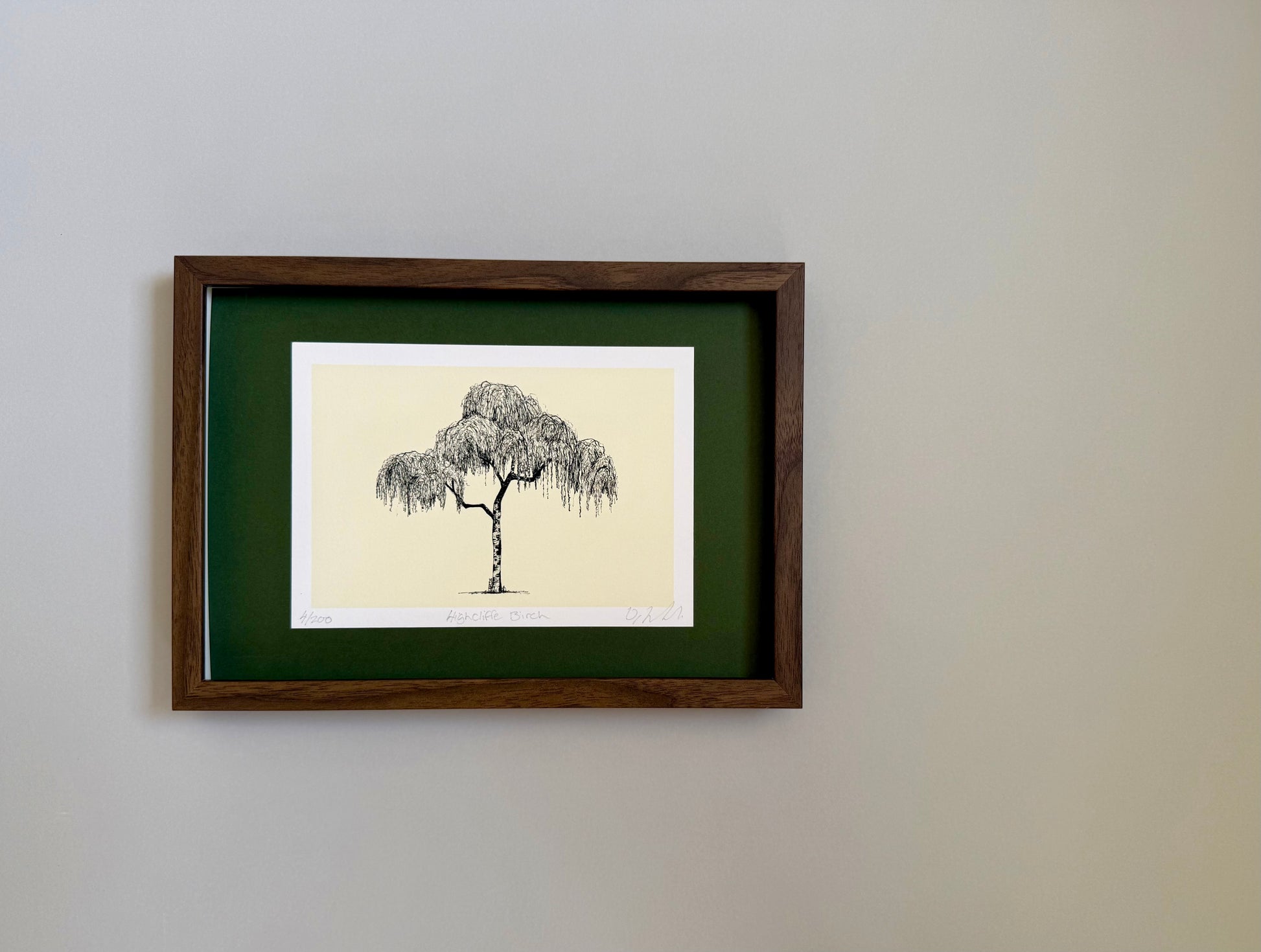A framed art print featuring a black ink sketch of a weeping tree on a cream background, surrounded by a white mat and a dark green mat board, enclosed in a dark brown wooden frame and mounted on a light gray wall.