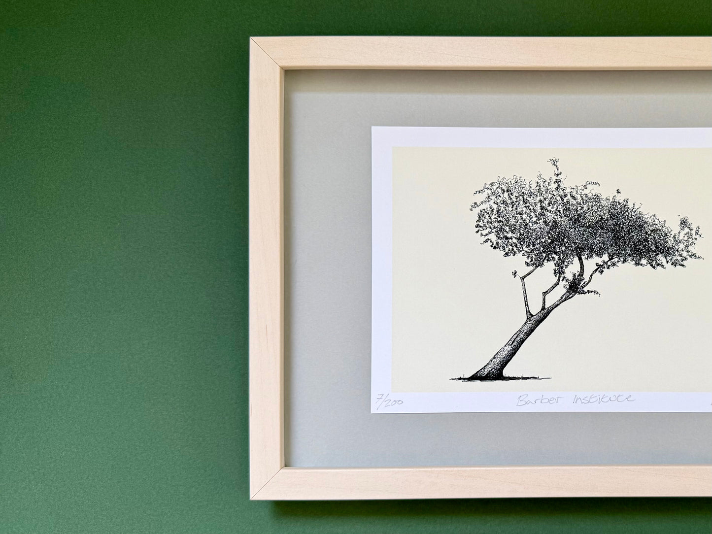 A framed art print featuring a black ink sketch of a leaning tree on a cream background, mounted with a gray mat inside a light natural wood frame against a dark green wall.