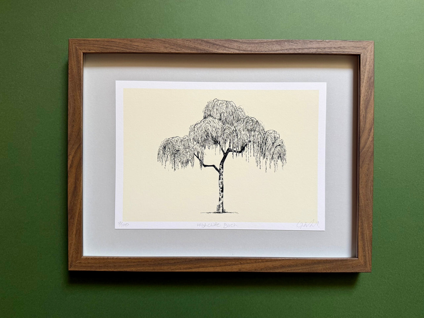 A framed art print featuring a black ink sketch of a weeping tree on a cream background, matted with white and enclosed in a medium brown wood frame, mounted on a textured dark green wall.