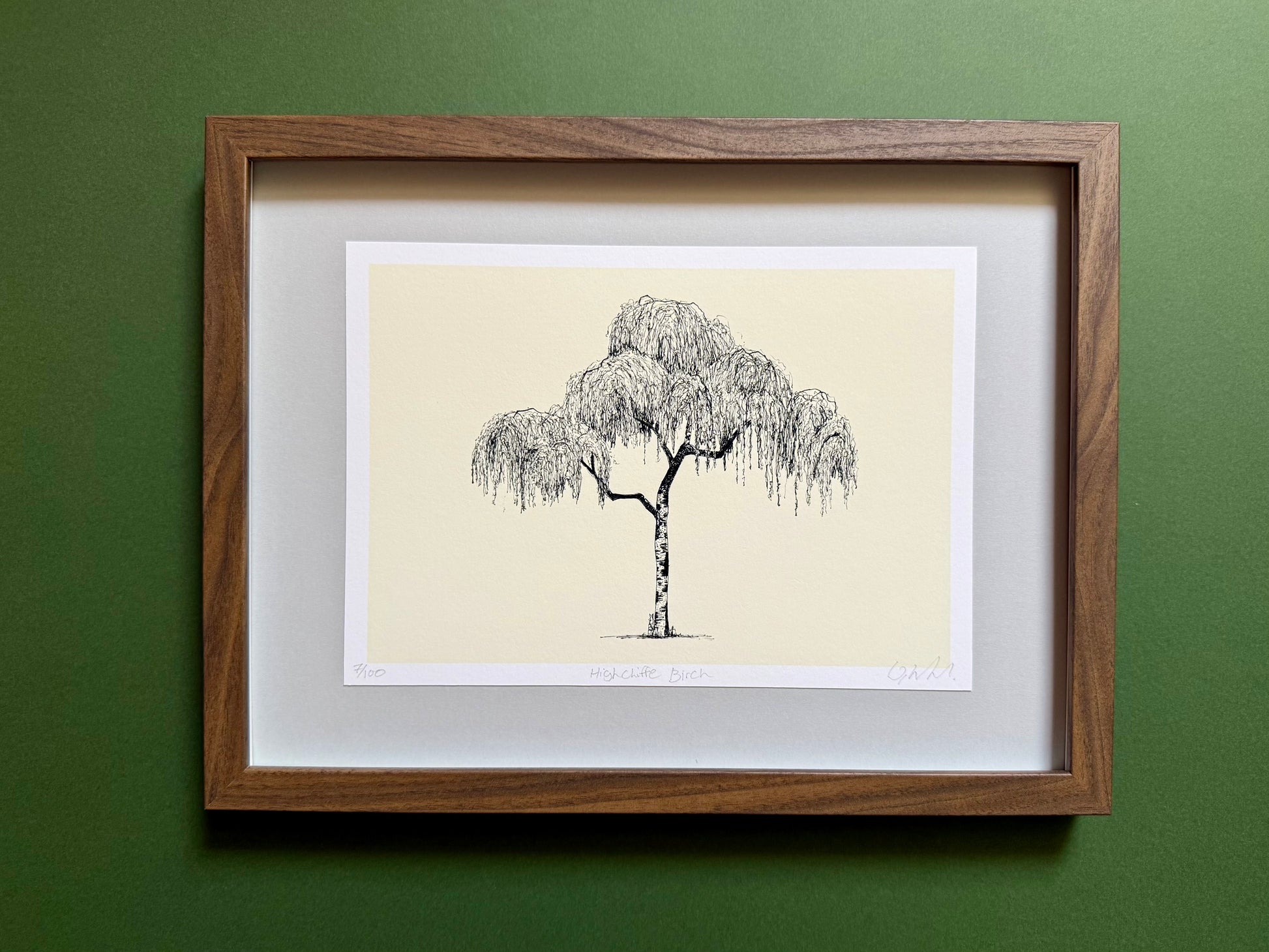 A framed art print featuring a black ink sketch of a weeping tree on a cream background, matted with white and enclosed in a medium brown wood frame, mounted on a textured dark green wall.