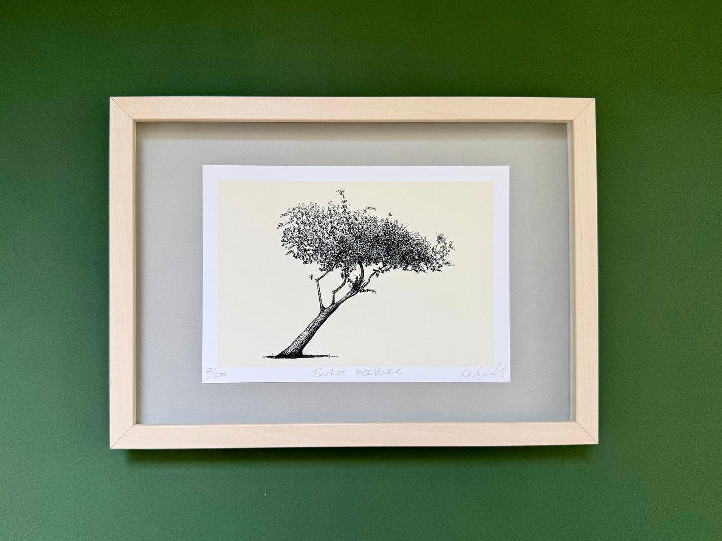 A framed art print featuring a black ink sketch of a leaning tree on a cream background, mounted with a gray mat in a light wood frame against a dark green wall.