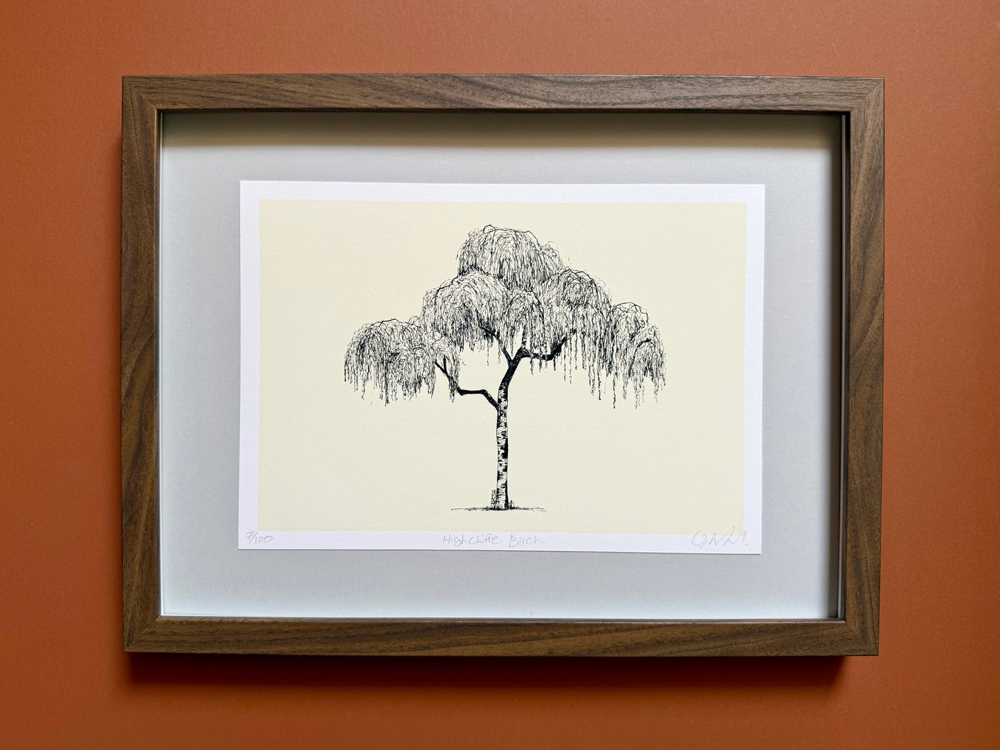 A framed art print featuring a black ink sketch of a weeping tree on a cream background, surrounded by a white mat and a dark brown wood frame, mounted on a terracotta-colored wall.