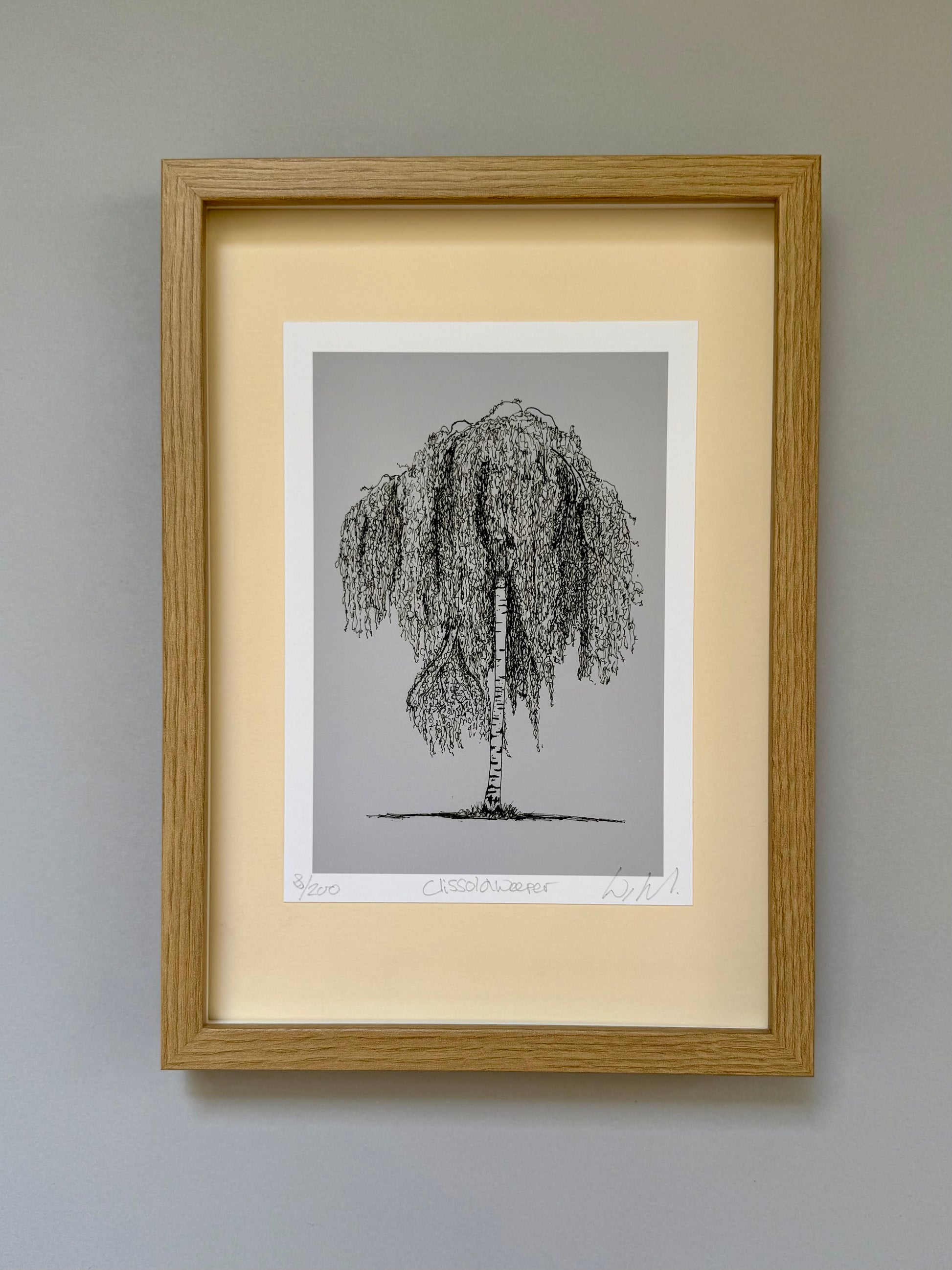 A framed art print featuring a black ink sketch of a weeping tree on a light gray background, set within a cream-colored mat and a natural oak frame, mounted on a neutral gray wall.