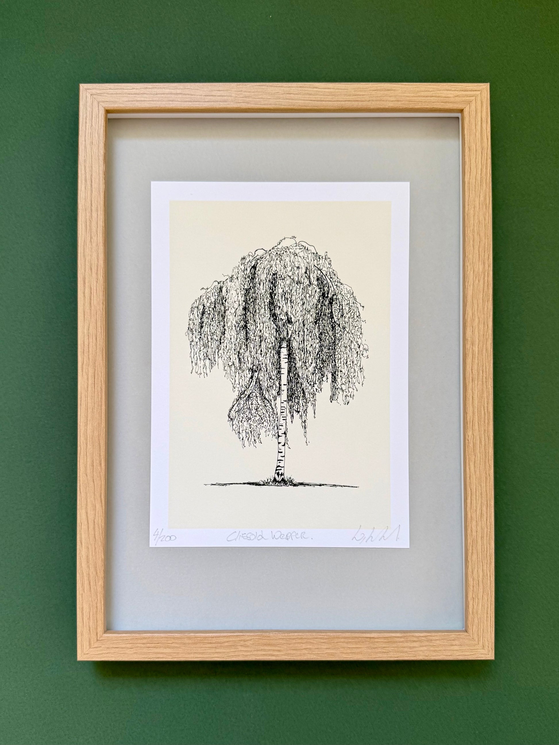 A framed black ink sketch of a weeping tree on a cream paper background, mounted with a white mat and a light natural wood frame against a dark green wall.