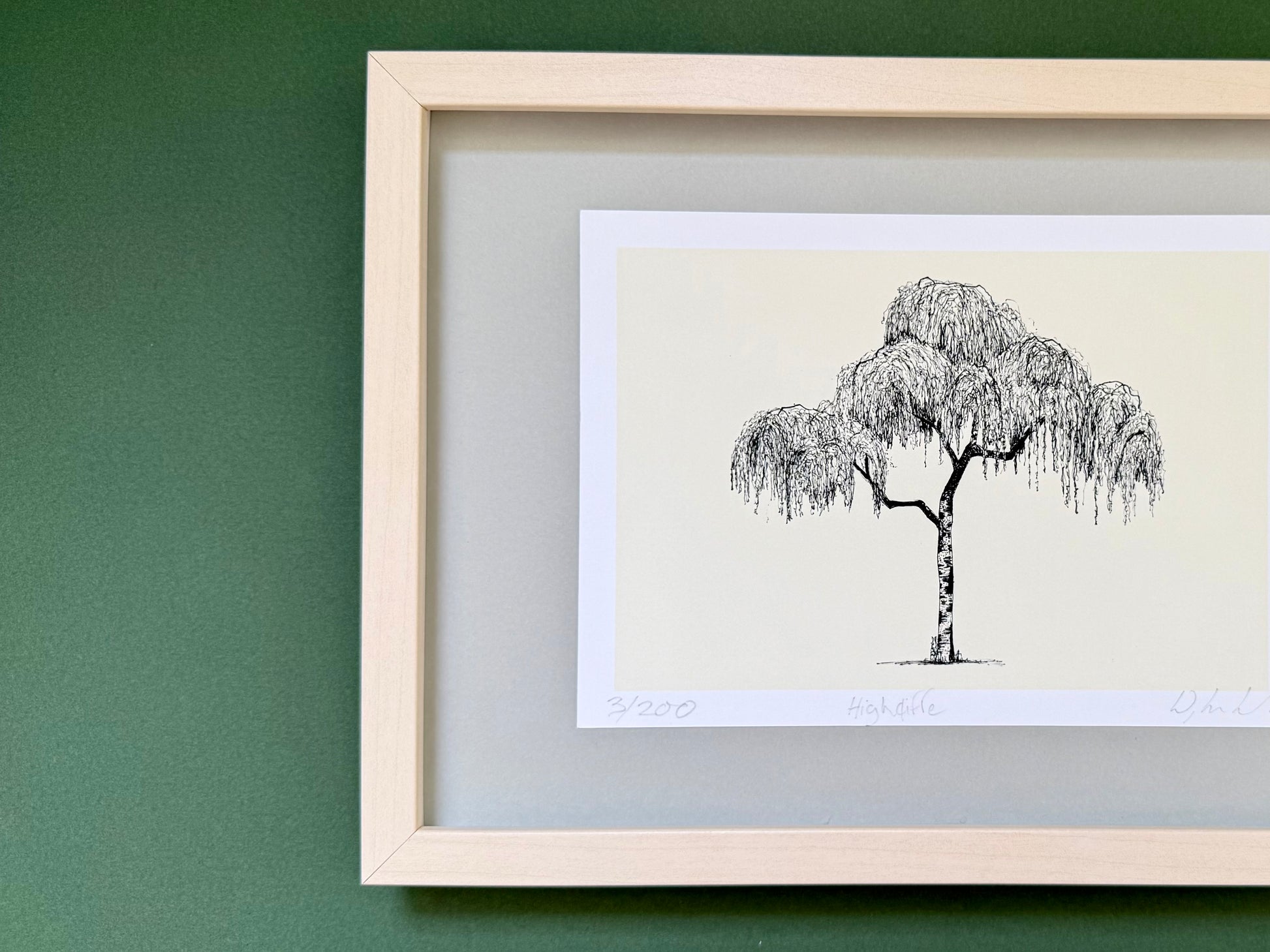 A framed art print featuring a black ink sketch of a weeping tree on an off-white background, mounted with a gray mat inside a light natural wood frame against a dark green wall.