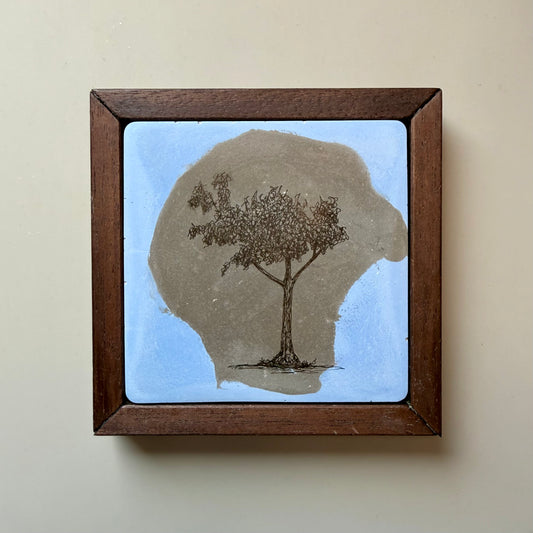 Ink drawing of a tree on a hand-cast cement tile pigmented in blue and natural grey, set in a dark wood frame against a warm off-white wall.