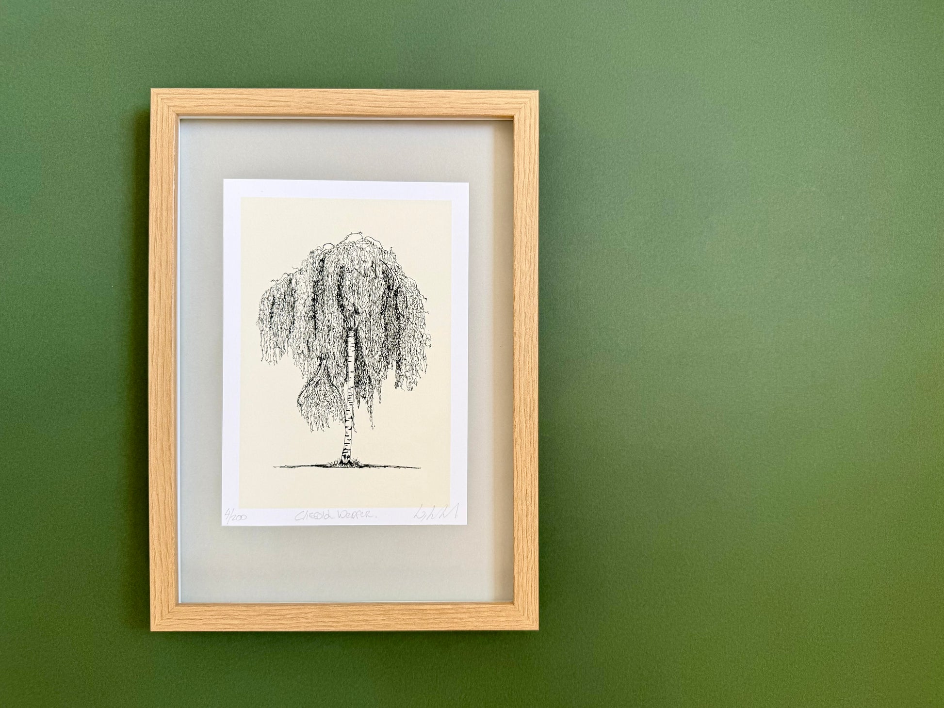 A framed art print featuring a black ink sketch of a weeping tree on a cream background, set in a light natural wood frame against a solid dark green wall.