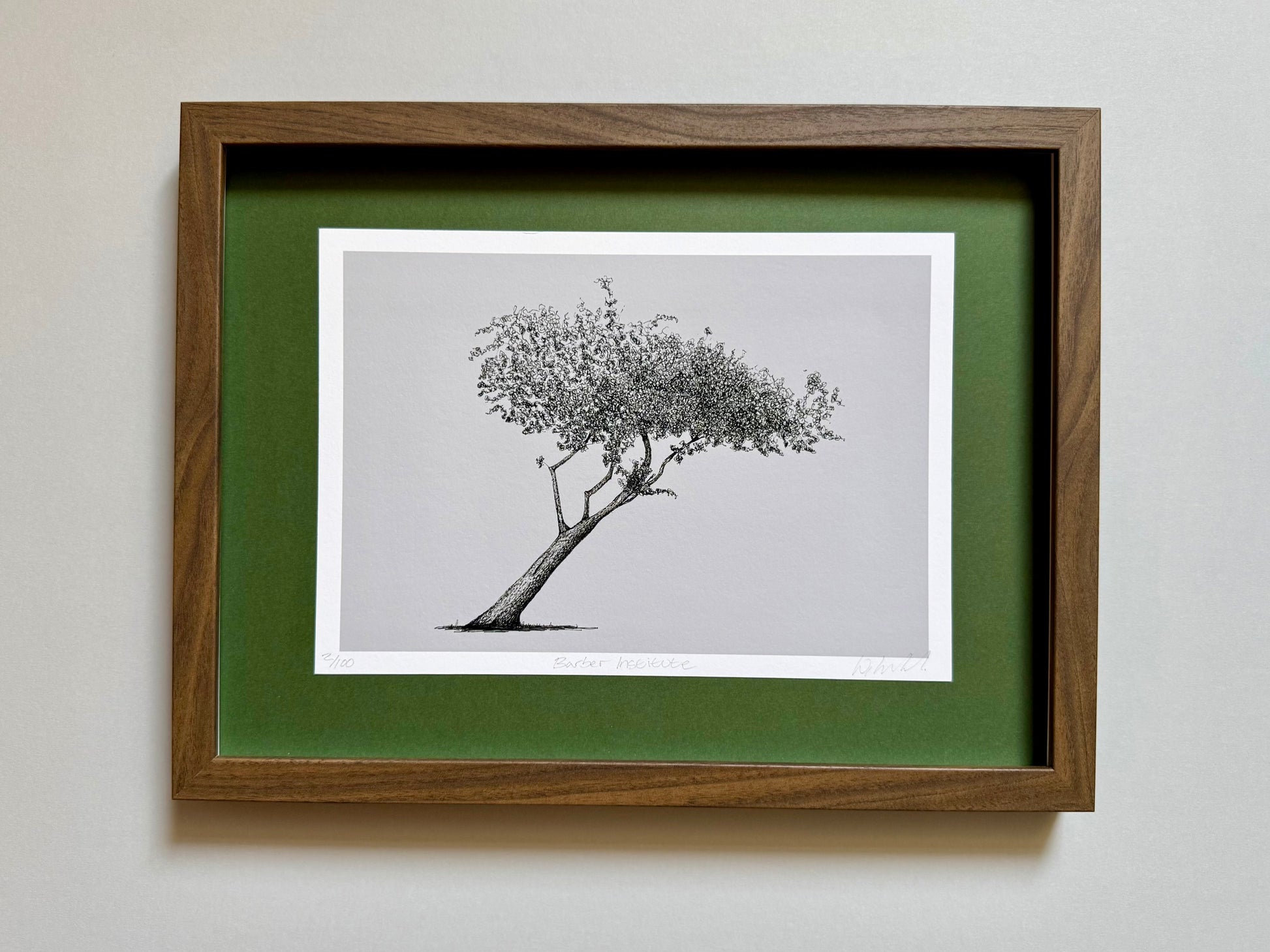 A framed art print featuring a black and white sketch of a leaning tree, set against a green mat and a medium brown wooden frame, mounted on a light gray wall.