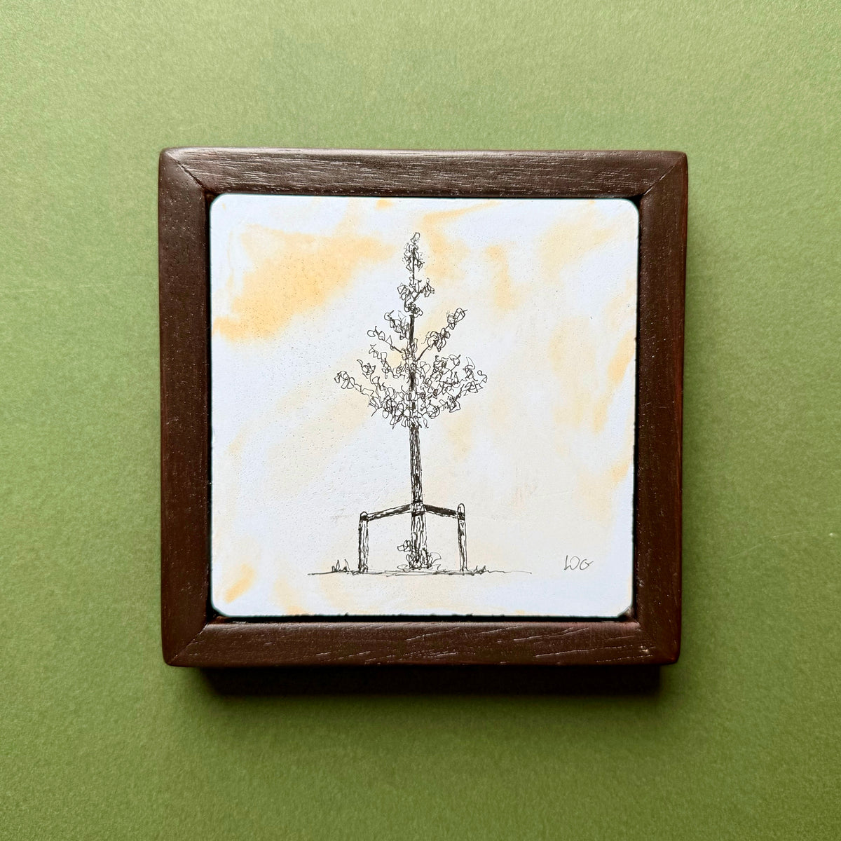 Original black ink drawing of a young Birmingham tree supported by wooden stakes, on a hand-cast Portland cement tile with soft orange and blue pigments. Presented in a dark wooden frame against a green background.