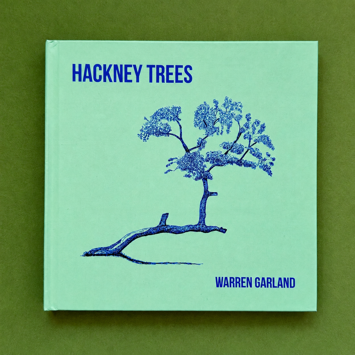 Hackney Trees second edition hardback artist book by Warren Garland, featuring a blue ink tree drawing on a mint-green cover, photographed against a green background.